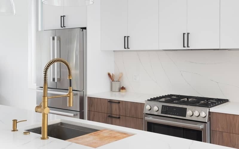 A kitchen detail with a gold faucet, white and wood cabinets, stainless steel appliances, and marble countertops