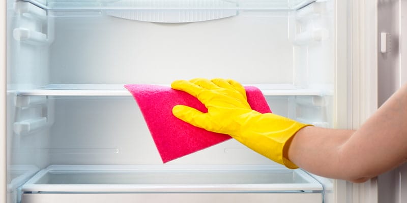 Woman's hand in yellow rubber protective glove cleaning white open empty refrigerator with pink cloth