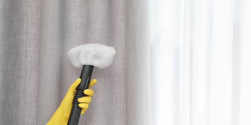 female hand with yellow gloves removing dust from curtain with steam cleaner indoors