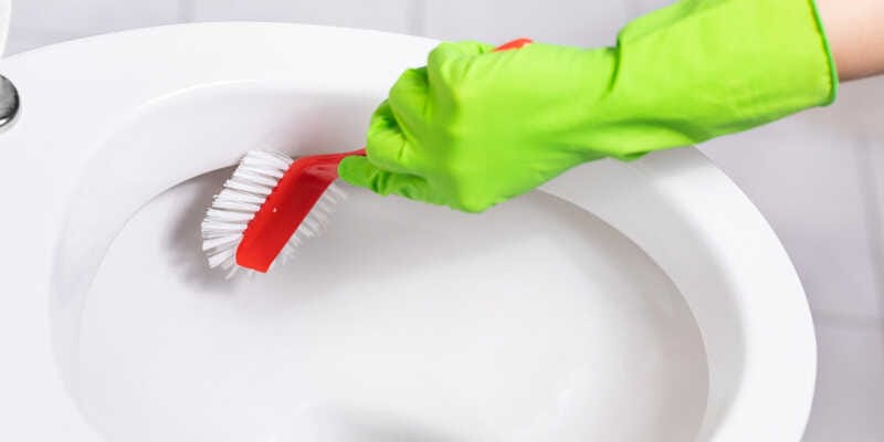 Woman with rubber glove is cleaning toilet bowl using brush