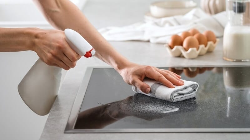 cropped shot of female hands cleaning electric stove top with cloth and spray bottle