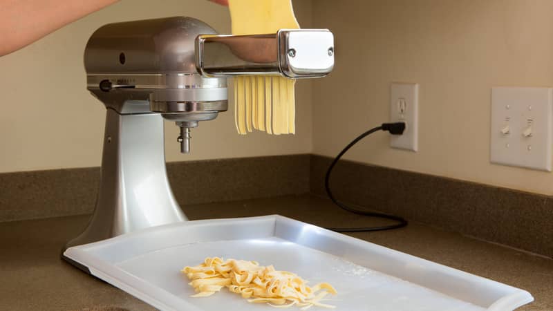 woman preparing homemade fettuccine pasta feeding the rolled sheets of dough through the electric cutting machine in the corner of a kitchen