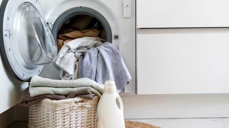 Front load washing machine with laundry spilling out, a basket of folded towels, and a bottle of detergent nearby