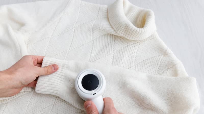 Man's hand holding a lint remover from a white acrylic or wool sweater.