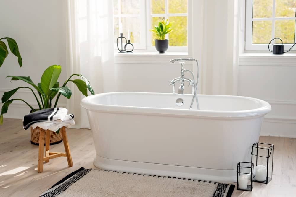 Stylish modern bathroom interior. Horizontal view of an empty free-standing bathtub on a wooden floor in a bright room against the backdrop of a large window and houseplants. Soft selective focus.