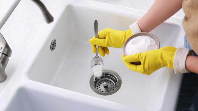 Woman in yellow gloves using baking soda to unclog sink drain