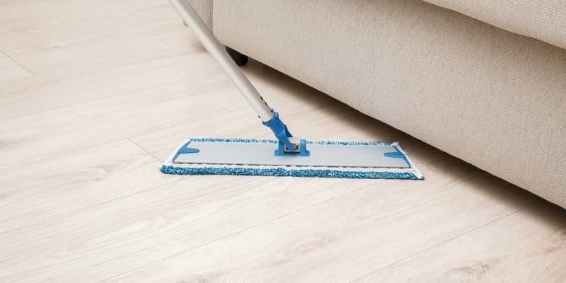 Microfiber mop pad with stick washing light laminate floor under beige sofa at living room. 