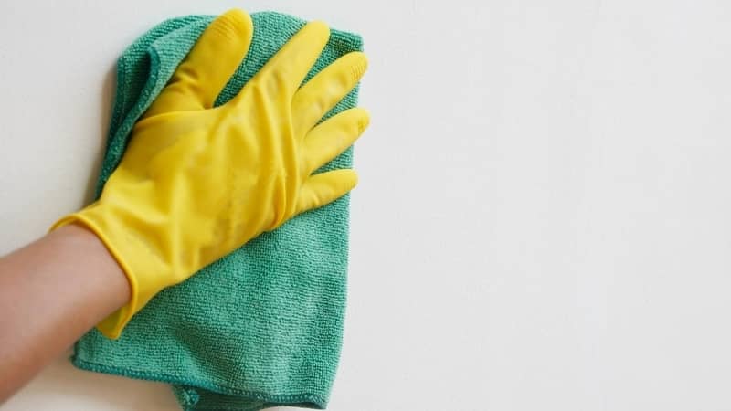 Hand wearing yellow rubber glove with green microfiber cloth cleaning wall.