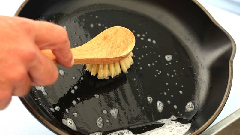 A hand holding a cleaning brush scrub and washing the dirty cast iron pan 