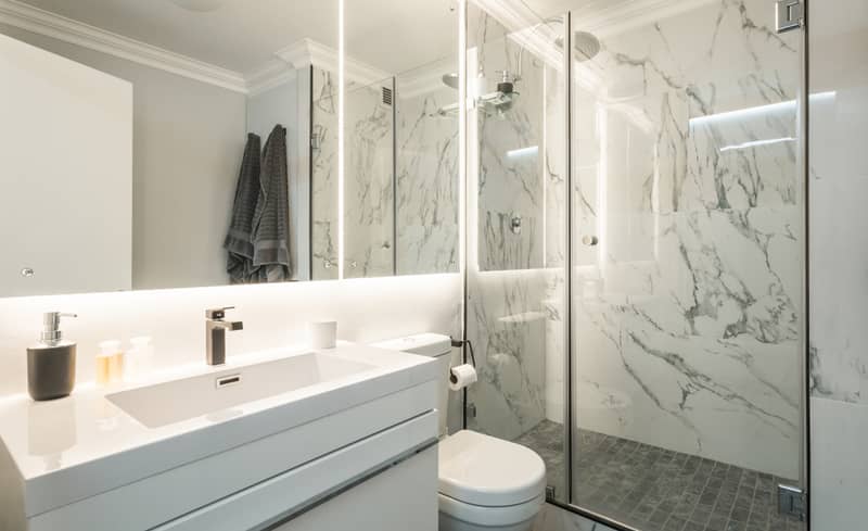 Modern, contemporary and brand new bathroom with marble walls and floor, white walls, cupboard and toilet, big back lit mirror