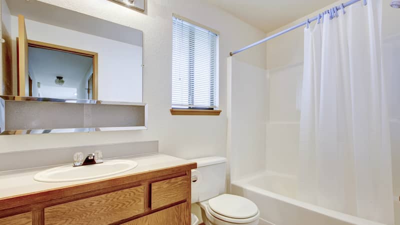 Bright bathroom with window, wooden washbasin cabinet, white toilet and tub with shower and shower liner. 