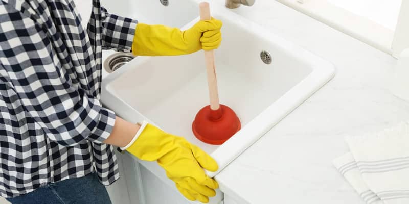 Woman using plunger to unclog sink drain in kitchen, closeup