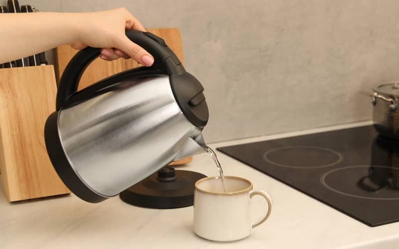 Woman pouring hot water from electric kettle into cup in kitchen, closeup