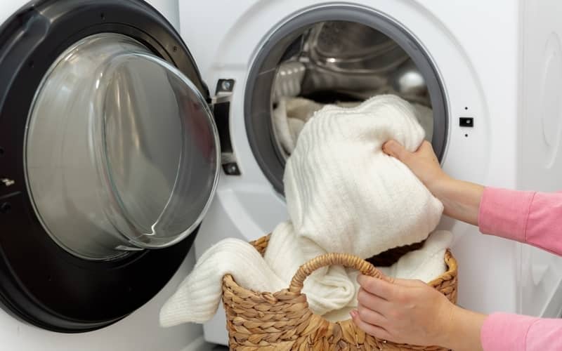 Woman putting white wool sweater and clothes into the drum of a front load washing machine